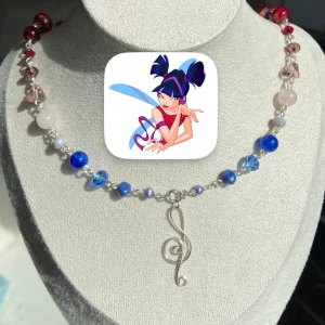 Färgglatt halsband med musikberlock - Unikt halsband med pärlor i blått, rött och lila samt en silverfärgad G-klav som hänge. Pärlorna är i olika former och storlekar, vilket ger ett lekfullt och personligt uttryck. Perfekt för dig som älskar musik och vill sticka ut med accessoarer. Inspirerat av karaktären Musa från tv-serien Winxclub.