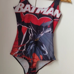 Batman body NY - Cool body med stort Batman-tryck i rött, svart och vitt. Snyggt motiv med Batman i action och loggan över bröstet. Klassisk modell med breda axelband och hög benskärning. Perfekt för dig som älskar superhjältar och vill sticka ut på stranden. Helt oanvänd!