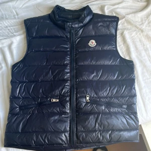 Mörkblå Moncler gui väst - Säljer en mörkblå dunväst från Moncler med klassisk quiltad design och dragkedja framtill. Västen har två fickor med dragkedja och Moncler-logga på bröstet. Perfekt för en sommarkväll, inget är sönder och nfc Scan fungerar toppen 