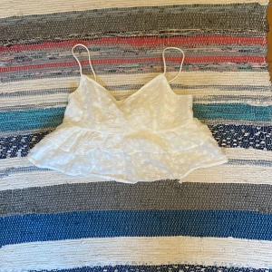 Vit volangig linnetopp med spets - Supersöt vit linnetopp med tunna axelband, volangig nederdel och broderade spetsdetaljer. Perfekt för en somrig look och lätt att matcha med jeans eller kjol. Luftig och feminin känsla.
