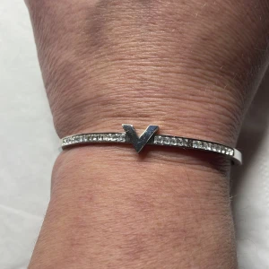 Silverfärgat armband med V-detalj och stenar - Stilrent armband i silverfärg med en rad gnistrande stenar och en tydlig V-formad detalj framtill. Armbandet är stelt och har en minimalistisk design som passar till många olika stilar.