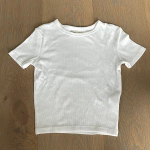 Vit ribbad t-shirt från Pull&Bear XS - En basic vit t-shirt från Pull&Bear i storlek XS. Modellen är ribbad med korta ärmar och rund halsringning. Perfekt att styla med jeans eller kjol för en clean look. Materialet är mjukt och stretchigt för extra komfort.