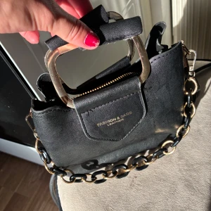 Svart handväska med kedjedetaljer - Snygg svart handväska från Fashion & Bags i skinnimitation med guldfärgade metalldetaljer och kraftig kedja som axelrem. Väskan har ett stilrent, fyrkantigt handtag och dragkedja upptill. Perfekt storlek för mobil, plånbok och småprylar.