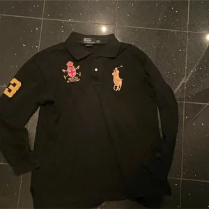 Svart långärmad pikétröja Polo Ralph Lauren - Svart långärmad pikétröja från Polo Ralph Lauren med klassisk krage, broderad gul logga på bröstet och broderat klubbmärke. Sitter som en liten medium. Skulle kunna passa S