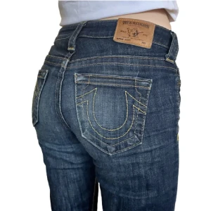 True Religion blå jeans med kontrastsöm - Säljer ett par klassiska blå jeans från True Religion med ikonisk gul kontrastsöm och tydlig logga på bakfickan. Jeansen har rak/flare passform, fem fickor och är tillverkade i robust denim. Snygga detaljer som läderpatch och amerikansk flagga på insidan.