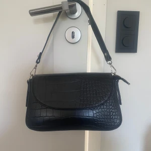 Svart axelväska från NA-KD - Snygg svart axelväska från NA-KD med croco-mönstrad yta och silvriga detaljer. Väskan är cirka 16x28 cm, skriv för mer specifika mått.