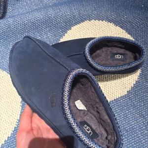 Blå mockatofflor från UGG - Mysiga blå tofflor från UGG i mocka med fluffigt foder och snyggt mönstrad kant i blått och beige. Perfekta för kalla dagar när du vill ha det extra bekvämt hemma. Klassisk UGG-logga på sidan och slitstark sula.