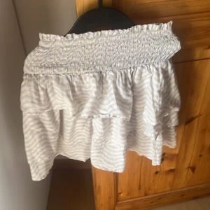 Randig volangkjol från H&M - Supersöt kort volangkjol från H&M i ljusblått och vitt med randigt mönster. Kjolen har bred smock i midjan och flera lager volanger som ger en flowy känsla. Perfekt för varma dagar när du vill ha en chill och fräsch look.