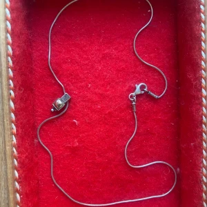 Silvrigt halsband med fyrkantig berlock - Ett smalt silvrigt halsband med en unik fyrkantig berlock. Berlocken har en liten infattad sten i mitten som ger en cool och stilren look. Kedjan är tunn och har ett klassiskt karbinlås. Perfekt för dig som gillar minimalistiska smycken med en twist.