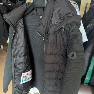 Svart Moncler dunjacka med ribbade ärmar - Snygg svart dunjacka från Moncler med quiltad front och huva. Ärmarna är ribbstickade för en modern look och jackan har dragkedja framtill. Perfekt för dig som vill ha en stilren och exklusiv jacka med streetkänsla.