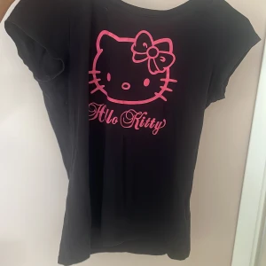 Svart Hello Kitty t-shirt - Svart t-shirt med korta ärmar och stort rosa Hello Kitty-tryck framtill. Klassisk rund halsringning och rak passform. Perfekt för dig som gillar en vintage cool stil och vill sticka ut med en ikonisk design. Vintage och finns därav inte att köpa längre! 