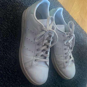 Adidas Stan Smith vita sneakers - Klassiska vita Adidas Stan Smith sneakers med gröna detaljer på hälen. Skorna har snörning, perforerade ränder på sidorna och är tillverkade i skinn. Perfekt till sommaren! Storlek 40 och 2/8