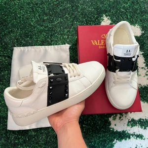 Valentino Rockstud Open  - Snygga Valentino Rockstud Open sneakers i vitt skinn med bred svart rem över vristen dekorerad med svarta nitar. Klassisk rund tå och platt sula, coola detaljer längs hälen och ikonisk design. Allt medföljer och en till valentino påse. OBS: inte orig1nal. Vid mer funderingar skriv
