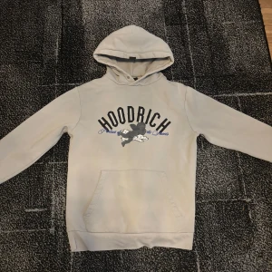 Beige Hoodie från Hoodrich med tryck - Snygg beige hoodie från Hoodrich med stor svart och blå logga framtill och detaljerat tryck på ryggen. Tröjan har huva, känguruficka och långa ärmar. Perfekt för dig som gillar streetwear och vill sticka ut med coola prints.