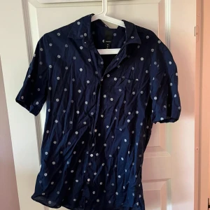 Mörkblå prickig skjorta H&M - Snygg mörkblå skjorta från H&M med vita prickar. Skjortan har korta ärmar, klassisk krage och knäppning framtill. Perfekt för dig som vill sticka ut lite extra med ett lekfullt mönster. Tillverkad i mjuk bomull för skön känsla.