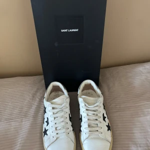 Saint Laurent sneakers med svarta stjärnor - Snygga vita sneakers från Saint Laurent i skinn med svarta stjärnor på , vita snören och beige sula. Perfekta för dig som vill sticka ut med en cool detalj men ändå hålla det stilrent. Hör av er vid minsta fråga. Priset är inte hugget i sten!😊
