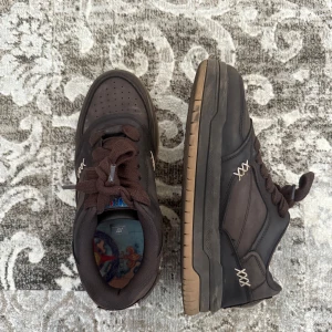 Bruna sneakers med One Piece-motiv - Unika bruna sneakers med grov sula och snygga detaljer i beige. Baksidan pryds av en stor 'Wanted' patch med Monkey D. Luffy från One Piece. Skorna har snörning och lågt skaft, samt coolt mönstrad innersula. Perfekta för dig som älskar anime och streetstyle.