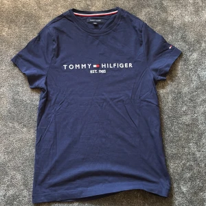 Marinblå t-shirt Tommy Hilfiger - Snygg marinblå t-shirt från Tommy Hilfiger med klassisk logga och texten 'EST. 1985' broderad på bröstet. T-shirten har rund hals, korta ärmar och liten flaggdetalj på ärmen. Perfekt för en clean och stilren look.
