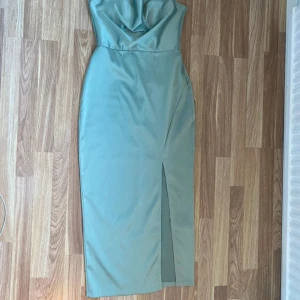Mintgrön långklänning med slits - Snygg mintgrön långklänning med draperad urringning och smala axelband. Klänningen har en figurnära passform och en slits framtill som ger en elegant touch. Materialet är glansigt och känns lyxigt, perfekt för speciella tillfällen.