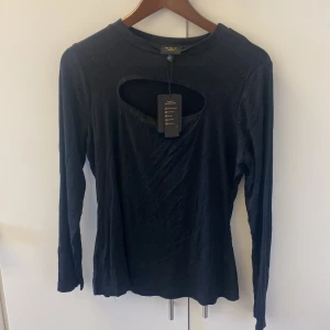 Svart långärmad topp med cut out MKF - Svart långärmad topp från M.K.F London med en snygg cut out-detalj vid bröstet. Modellen är figurnära och ribbad, vilket ger en trendig och stilren look. Perfekt för dig som vill ha något enkelt men ändå med en twist.