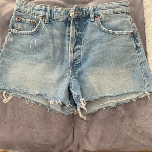 Ljusblå jeansshorts från Zara - Ljusblå jeansshorts från Zara med hög midja, råa fransiga kanter och klassisk femficksdesign. Shortsen har knäppning med knappar framtill och är tillverkade i denim. Perfekta för en chill sommarlook.