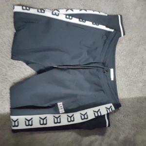 Svarta Better Bodies byxor med logga - Svarta byxor från Better Bodies med vita sidor och svart logotyp längs benen. Tight passform och snörning i midjan. Perfekta för träning och sportiga aktiviteter. Materialet är stretchigt och känns sportigt.