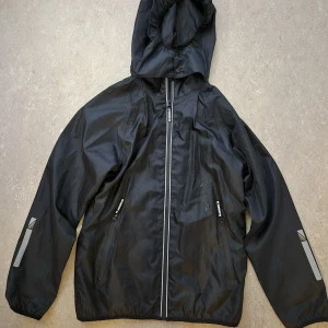 Regnjacka - Säljer en svart regnjacka från Mywear med huva och dragkedja. Jackan har långa ärmar och reflexdetaljer på ärmarna och ryggen. Finns fläck på framsidan. Perfekt för regniga dagar och utomhusaktiviteter.