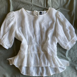 Vit blus med volang - Superfin vit blus med puffiga korta ärmar, volang nertill och knytning i ryggen. Tillverkad i ett luftigt linnematerial som ger en avslappnad men ändå söt look.