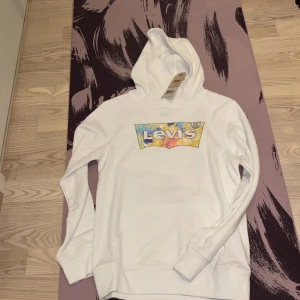 Vit hoodie från Levi's med färgglad logga - Säljer en vit hoodie från Levi's med en stor, färgglad batik-inspirerad logga på bröstet. Tröjan har huva, lång ärm och en klassisk känguruficka framtill. Perfekt för dig som gillar streetstyle och vill sticka ut lite extra.