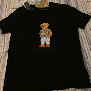 Svart Polo Bear t-shirt från Ralph Lauren - Svart t-shirt från Ralph Lauren med ikoniska Polo Bear-trycket på bröstet. Björnen har färgglad skjorta, vita byxor och solbrillor. Klassisk passform och rund hals. Perfekt statement-plagg för dig som gillar streetstyle och snygga prints. Säljer en helt ny polo t-shirt som ja fick i present men tyvärr så sitter den tight på mig. Priset kan diskuteras! och skriv till mig om frågor och funderingar! Obss den e slim fitt så den passar size M och S också