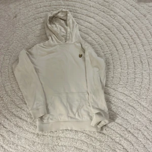 Vit hoodie från Lyle & Scott Junior - Vit hoodie från Lyle & Scott Junior med klassisk gul fågel-logga på bröstet. Tröjan har huva, magficka och långa ärmar. Perfekt för en chill och stilren look. Tillverkad i mjuk bomull för extra komfort.