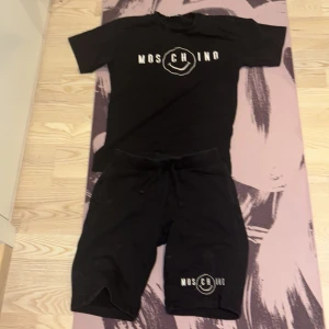 Svart Moschino t-shirt med tryck och shorts  - Svart t-shirt från Moschino med vitt tryck och shorts, logga framtill. Klassisk rund hals och korta ärmar. Perfekt för dig som gillar streetwear och vill ha en stilren look med en twist. Skön passform och enkel att matcha med andra plagg.