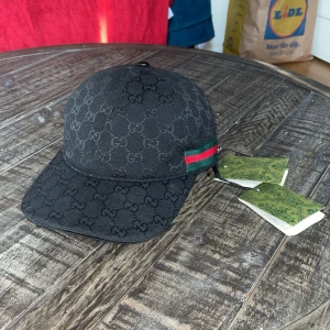 Svart GG keps med GG-mönster - Snygg svart keps från GG med klassiskt GG-monogram över hela kepsen. På sidan finns en ikonisk grön och röd rand. Justerbar rem baktill och etiketter kvar. Perfekt för dig som vill ha en exklusiv streetstyle-look.