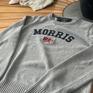 Grå Morris tröja med brittisk flagga - Snygg grå tröja från Morris med broderad brittisk flagga och tydlig logga framtill. Tröjan har ribbade muddar vid ärmslut och nederkant samt rund halsringning. Perfekt för dig som gillar klassisk och stilren design.