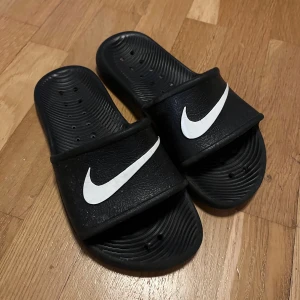 Svarta Nike sandaler med vit logga - Svarta sandaler från Nike med bred rem och stor vit Swoosh-logga. Skorna har öppen tå och platt sula med räfflad insida för extra grepp. Perfekta för sommaren eller efter träningen.