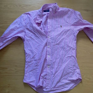 Rosa- och vitrandig skjorta från Ralph Lauren - Säljer en klassisk rosa- och vitrandig skjorta från Ralph Lauren med broderad logga på bröstet. Skjortan har lång ärm, knappar hela vägen och en snygg krage. Perfekt för dig som gillar preppy stil och vill sticka ut lite extra. Passar även M