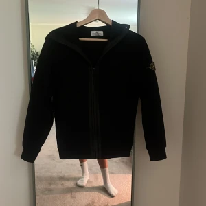 Svart hoodiejacka från Stone Island - Svart hoodiejacka från Stone Island med klassisk logga på ärmen. Jackan har dragkedja framtill, huva och är tillverkad i mjuk polyester med fleece på insidan. Perfekt för dig som gillar en clean look. Säljer för att jag har växt ut den. Tveka inte att höra av dig vid frågor!
