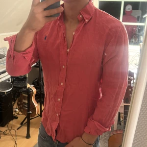 Rosa skjorta från Ralph Lauren - Säljer en 100% linen snygg rosa skjorta från Ralph Lauren med klassisk button-down krage och blå broderad logga på bröstet. Skjortan har långa ärmar, vita knappar och är tillverkad i bomull. Perfekt för dig som vill ha en stilren och tidlös look.