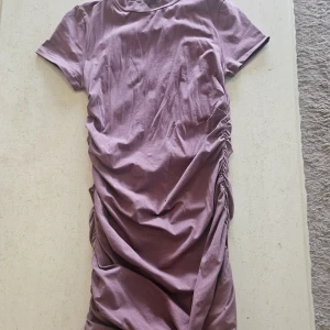 Ljuslila kortklänning från NLY Trend - Kort klänning i ljuslila från NLY Trend. Modellen har korta ärmar, rund hals och snygg rynkdetalj längs sidorna som ger en cool draperad effekt. Tillverkad i mjuk bomull för en bekväm känsla hela dagen.