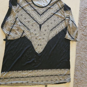 Svart och guldig topp från Xin Lai - Snygg svart topp från Xin Lai med guldigt paisleymönster och korta ärmar. T-shirten har rund halsringning och ett detaljerat tryck både fram och på ärmarna. Perfekt för dig som gillar unika prints och vill sticka ut lite extra.