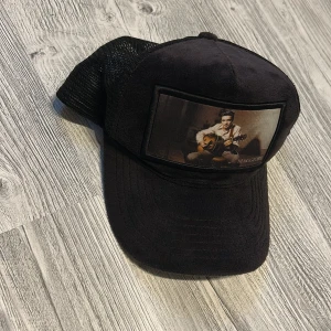 Svart keps Maggiore med Elvis Presley  - Svart keps från Maggiore med mesh baktill och justerbar snapback. Framtill finns en stor patch med motiv av en person som spelar gitarr. Kepsen har broderad logga på insidan och är tillverkad i syntetmaterial med tygpanel framtill.