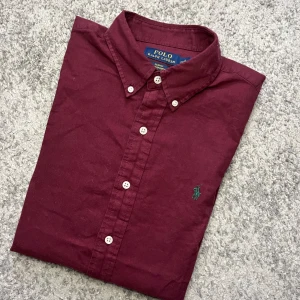 Vinröd skjorta från Polo Ralph Lauren - Säljer en vinröd långärmad skjorta från Polo Ralph Lauren i slim fit. Skjortan har klassisk krage, vita knappar och grönt broderat logotyp på bröstet. Tillverkad i bomull och passar perfekt till dig som gillar stilrena plagg.