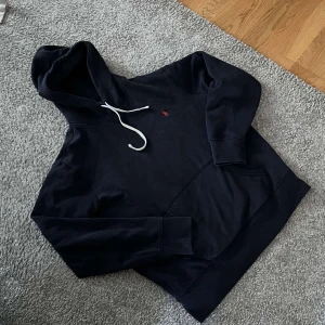 Marinblå hoodie från Ralph Lauren - Marinblå hoodie från Ralph Lauren med vit snörning i huvan och klassisk röd logga broderad på bröstet. Tröjan har känguruficka och långa ärmar, perfekt för en chill och stilren look.