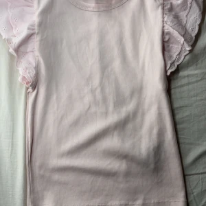 Ljusrosa topp med volangärm - Supersöt ljusrosa topp från Gina Tricot med ribbad struktur och breda volangärmar i broderad bomull. Perfekt för dig som gillar romantiska detaljer och en feminin look. Toppen är ärmlös med volang och har rund halsringning.