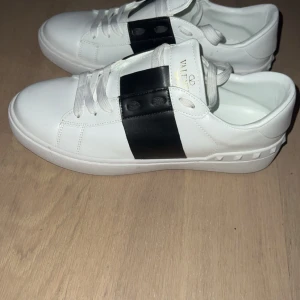Valentino Garavani Open sneakers vit/svart - Snygga Valentino Garavani Open sneakers i vitt skinn med bred svart läderdetalj över snörningen. Klassisk rund tå, platt sula och vita skosnören. Guldtext på plösen och insidan. Perfekta för dig som gillar stilrena och lyxiga sneakers.