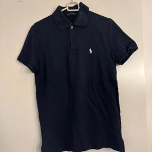 Marinblå pikétröja från Ralph Lauren - Klassisk marinblå pikétröja från Ralph Lauren med vit broderad logga på bröstet. Tröjan har korta ärmar, krage och knappar framtill. Tillverkad i mjuk bomull som känns skön mot huden. Perfekt för en clean och stilren look.