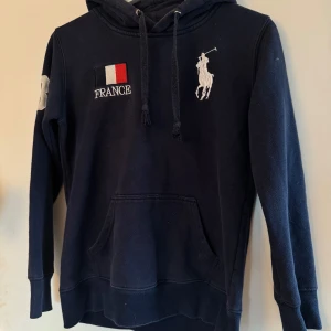 Mörkblå hoodie Polo Ralph Lauren France - Mörkblå hoodie från Polo Ralph Lauren med broderad fransk flagga och texten FRANCE på bröstet, stor vit polospelare-logga och nummer 3 på ärmen. Känguruficka framtill, snörning i huvan och broderade detaljer på ryggen. Perfekt för dig som gillar sportig och klassisk stil.
