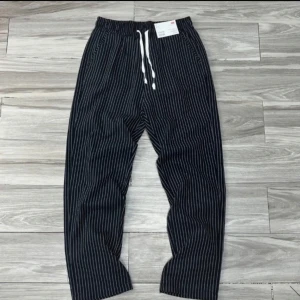 pinstripe pants - Snygga svarta byxor från Uniqlo med vita tunna ränder och elastisk midja med vit snörning. Byxorna har avslappnad passform och raka ben, perfekta för en chill och stilren look. Tillverkade i mjukt bomullsmaterial för extra komfort.