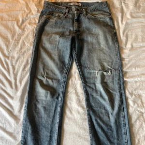 Lee Premium Regular Straight Leg jeans - Ett par klassiska blå jeans från Lee Premium med rak passform och regular midja. Jeansen har slitningar och en trasig detalj på ena benet för en avslappnad look. Fickor fram och bak, samt traditionell knapp och dragkedja. Perfekta för en casual stil.