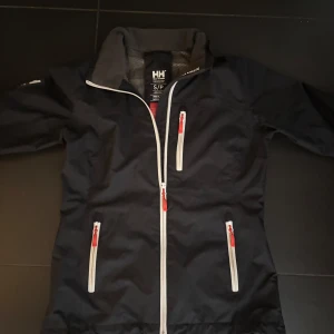 Svart vindjacka från Helly Hansen - Säljer en svart vindjacka från Helly Hansen med Helly Tech Protection. Jackan har dragkedja framtill och flera praktiska fickor med vita och röda detaljer. Perfekt för blåsiga dagar och utomhusaktiviteter.